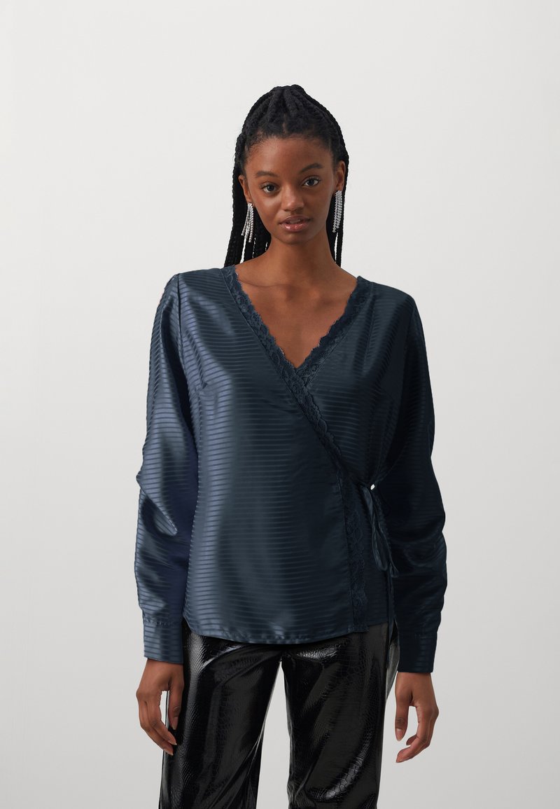 Marine satin omslagbluse med V-hals, prydet med blondepynt og lange ermer. Båret med skinnende svarte bukser som viser en glatt tekstur.