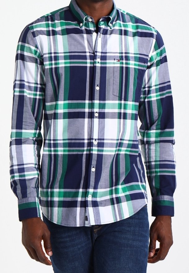 Homme portant une chemise à manches longues boutonnée avec un motif à carreaux bleu marine, vert, blanc et gris, et un jean bleu.