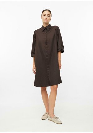 Vrouw die een losse, zwarte blousejurk tot op de knie draagt met opgerolde mouwen en beige casual schoenen tegen een effen witte achtergrond.