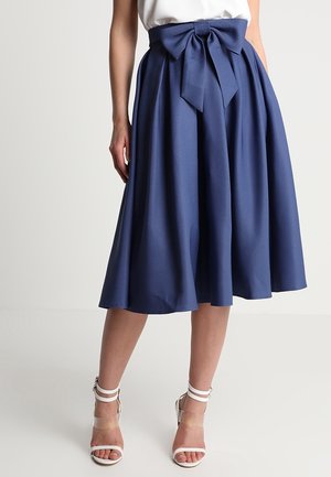 Femme portant une jupe midi taille haute bleu marine avec un grand nœud à l'avant, associée à des sandales blanches à talons hauts et à lanières.