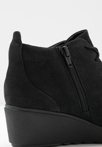 Bottine à talon compensé noir avec fermeture éclair latérale et détail à lacets, cuir texturé et coutures cousues sur un fond uni.