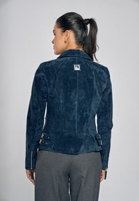 Donkerblauwe jas van nepsuède met een getailleerde pasvorm, voorzien van een nekkraag, decoratieve gespen bij de taille en ritssluitingen aan de mouwen.