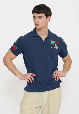 Giovane uomo con capelli ricci scuri che indossa una polo blu navy con palma e testo "Out of Office", pantaloni beige, in piedi con le mani sui fianchi.