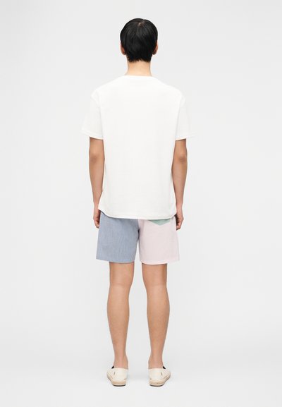 Άτομο που στέκεται γυρισμένο προς τα πίσω, φοράει απλό λευκό T-shirt, σορτς με μπλοκ χρωμάτων σε μπλε και ροζ ρίγες, και λευκά παπούτσια χωρίς κορδόνια.