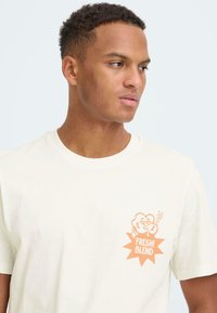 Crèmekleurig katoenen t-shirt met een perzikkleurige cartoonafbeelding en de tekst "FRESH! BLEND" op de linkerborst. Ronde halslijn, korte mouwen.