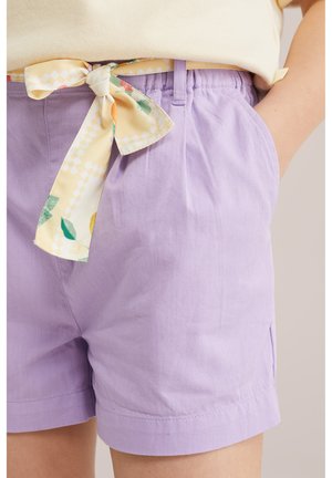 Svetlo vijolični bombažni kratke hlače z elastičnim pasom in rumenim cvetličnim pasom iz blaga, zavezanim v pentljo, nošene s svetlo rumeno majico.
