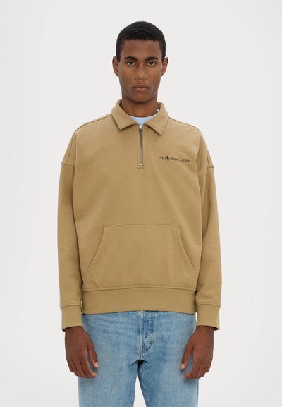 Brown zip-neck pullover sweatshirt with a front pocket and ribbed hem. Features a logo on the left chest. Worn with light blue jeans.