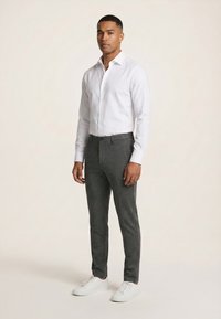 Homme portant une chemise blanche ajustée, un pantalon gris slim et des baskets blanches, debout devant un fond clair uni.