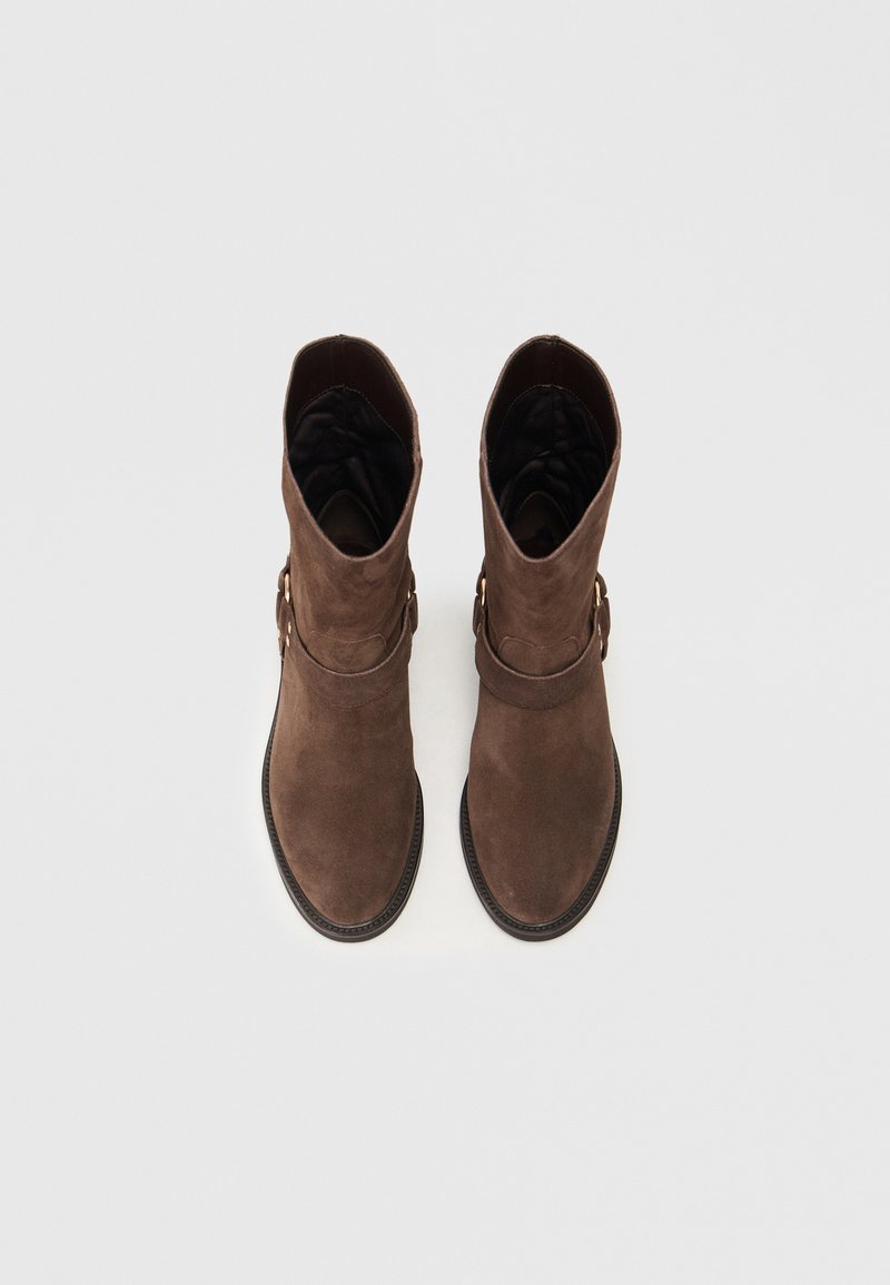 Braune Wildleder-Stiefeletten mit glatter Textur, runder Zehenform und einem dekorativen Riemenakzent. Innenfutter am oberen Rand sichtbar.
