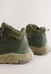 Žalios spalvos žygio batai, pagaminti iš Cordura audinio ir gumos, su paminkštintu apykakle, reljefine paviršiaus tekstūra ir stambiu smėlio spalvos padu su sukibimo raštais.