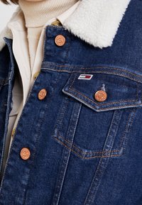 Närbild av en denimjacka med vit fårskinnskrage, kopparknappar och en liten röd, vit och blå logotyp på bröstfickan.