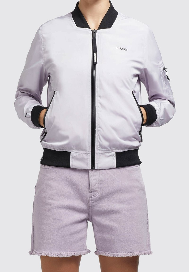 Personne portant une veste bomber violet clair avec bordure noire et poches zippées, associée à un short en jean violet clair effiloché.