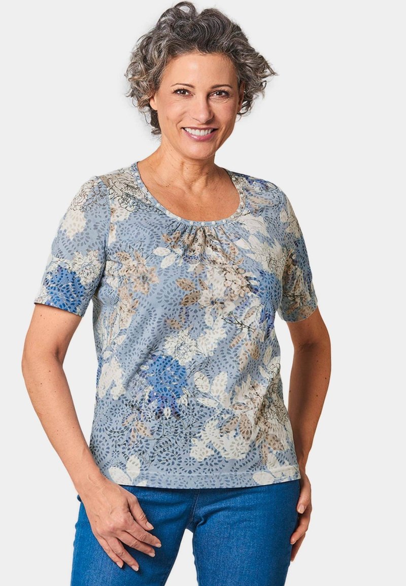 Kurzärmliges Oberteil in Hellblau mit einem floralen Muster in Weiß- und Blautönen, gerafftem Ausschnitt und weichem Stoff. Kombiniert mit blauen Jeans.