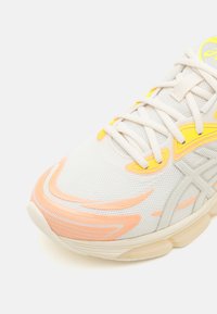 Sneaker bianca con tomaia in rete, caratterizzata da accenti gialli e linee di design arancioni. La suola è spessa e di colore crema, con grip testurizzato.