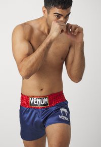 Pantalones cortos de boxeo de satén azul con una cintura elástica roja, que presentan un logo blanco y el texto "VENUM" en negro, diseñados para uso atlético.