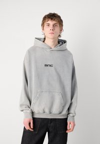 Sudadera de algodón gris con un amplio bolsillo frontal, capucha con cordón y logotipo negro en el pecho. Presenta puños y dobladillo de canalé.