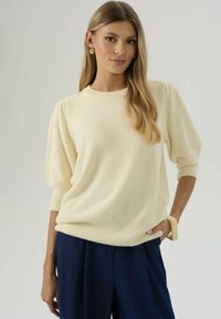 Jeune femme aux longs cheveux blonds portant un pull ivoire à manches bouffantes, un pantalon large bleu marine, des boucles d'oreilles en or et un bracelet beige épais.