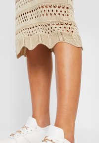 Beige Strickrock mit offenen Mustern und gewelltem Saum, kombiniert mit weißen Sneakers mit goldenen Akzenten und Schnürsenkeln, die eine glatte Textur zeigen.