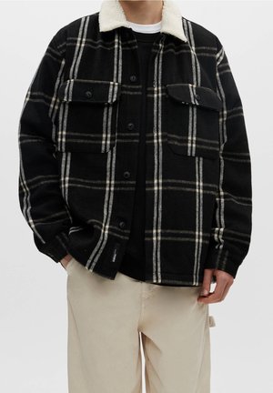 Man die een zwart-wit geruite jas met knopen draagt, met een witte fleece kraag, een zwart shirt eronder en beige broek, staand met handen in de zakken.