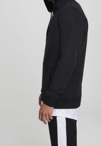 Svart hoodie i mjukt material, med dragkedja vid halsen, framficka och ribbade ärmavslut. Bärs över en vit lagertröja och svarta byxor.