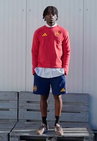 Crvena Adidas majica dugih rukava sa žutim detaljima, uparena s tamnoplavim šortsevima s logotipom. Model nosi smeđe cipele i crne čarape, stojeći na drvenoj platformi.