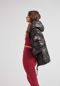 Svart pufferjacka med en glänsande yta, fastsydd huva och en rosa accentpatch. Bärs över en röd ribbad crop top och matchande leggings.