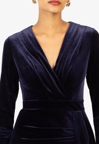 Marineblauwe fluwelen jurk met een diepe V-hals, lange mouwen en een aansluitende taille met geplooide accenten. Zachte textuur en luxe afwerking.
