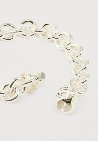 Sudraba krāsas rokassprādze no pulētiem metāla posmiem, ar apaļu aizdari ar gravējumiem. Gluds tekstūras un savstarpēji savienots dizains.