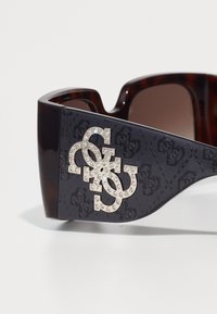 Gafas de sol de carey con un lateral de plástico oscuro que presenta un diseño de logo en relieve adornado con rhinestones. Lentes de tonalidad marrón.