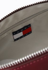 Burgundy Tommy Jeans -laukun sisäpuoli, jossa on vaalea vuori, vetoketjullinen tasku ja etiketti, jossa on sinisiä, punaisia ja valkoisia graafisia elementtejä.