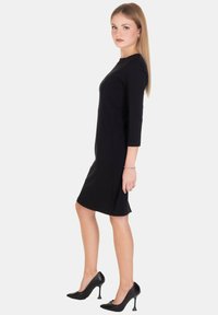 Abito aderente nero con maniche a tre quarti, orlo al ginocchio e texture liscia, abbinato a scarpe nere con tacco alto. Design semplice.