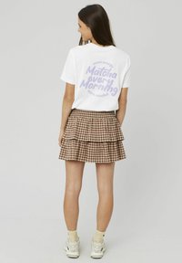 Jeune femme aux longs cheveux bruns portant un t-shirt blanc avec un texte violet et une jupe en tiers à carreaux bruns, se tenant de dos.
