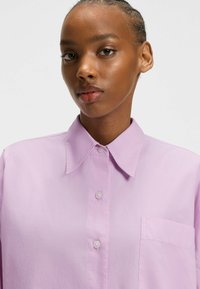 Camicia button-up lavanda con colletto a punta, tasca frontale e texture liscia. Presenta bottoni trasparenti e un design dal fit rilassato.