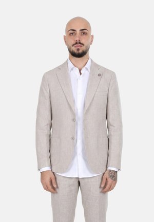 Uomo calvo con barba che indossa una giacca da completo beige chiaro a due bottoni e pantaloni abbinati sopra una camicia bianca, in piedi davanti a uno sfondo semplice.