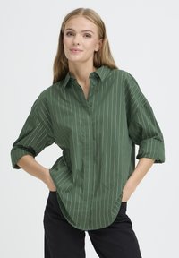 Camicia di cotone verde con righe verticali bianche, colletto button-down, maniche arrotolate e orlo curvo.