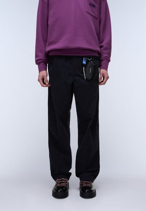 Osoba ubrana w fioletową bluzę, czarne spodnie z małą saszetką przypiętą oraz czarne buty z różowymi sznurówkami, stojąca na neutralnym tle.