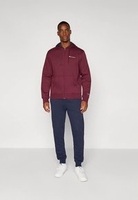 Donkerrode capuchontrui met rits, trekkoorden en voorzakken, gecombineerd met marineblauwe joggingbroek en witte sneakers. Gladde textuur, casual pasvorm.