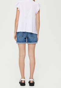 Hellblaues Oberteil mit Rüschenshirt und kurzen Ärmeln, kombiniert mit blauen Denim-Shorts mit umgeschlagenen Säumen und Gesäßtaschen, getragen mit weißen Sandalen.