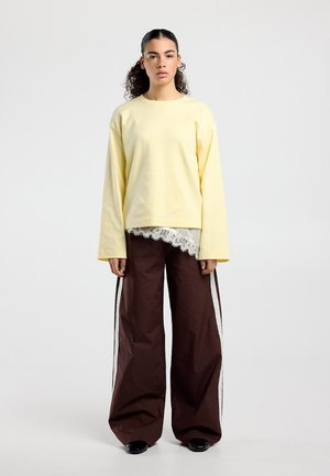 Femme aux cheveux bouclés portant un haut à manches longues jaune pâle, un sous-vêtement blanc orné de dentelle, un pantalon large marron avec des bandes blanches sur les côtés, et des chaussures noires.