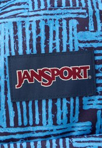 Náplasti s logem Jansport přišité na modrý materiál s tmavě modrým abstraktním proužkovaným vzorem.