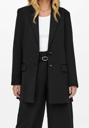 Kvinna klädd i en svart skräddarsydd blazer över en vit topp, tillsammans med höga svarta vida byxor och ett svart bälte med silverspänne.