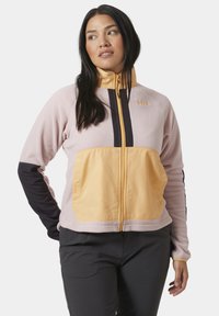 Chaqueta de forro polar en rosa claro con un bolsillo en el pecho amarillo, mangas negras y cremallera. Presenta franjas horizontales en contraste y detalle del logo.