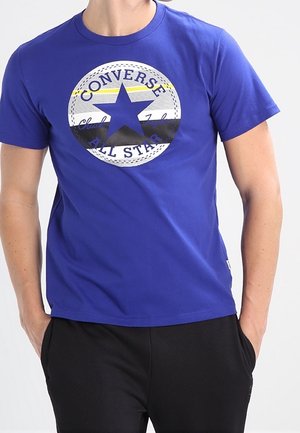 Sininen puuvillainen t-paita, jossa suuri musta tähtikuosi ja "Converse All Star" -teksti ympyrämuodostelmassa. Lyhyet hihat ja pyöreä pääntie.