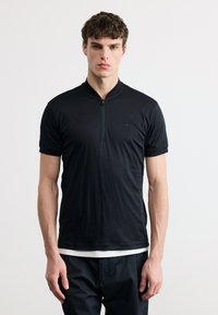 Νεαρός άνδρας που φοράει μαύρο κοντομάνικο πουκάμισο με φερμουάρ πάνω από λευκό εσώρουχο, στέκεται μπροστά σε ένα απλό ανοιχτό φόντο.