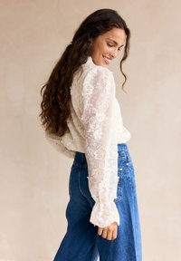 Blusa di pizzo con motivi floreali e maniche lunghe a campana, abbinata a jeans in denim a vita alta. Colore crema chiaro, texture trasparente.