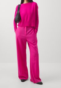 Fuksia neulepusero, röyhelöhattu; fuksia leveälahkeiset housut, joissa on taskut; musta käsilaukku; tasapohjaiset mustat kengät, joissa on kärjet.