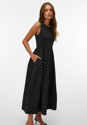 Femme portant une robe noire sans manches avec un haut ajusté et une longue jupe à pois, debout avec une main dans la poche et portant des talons noirs.