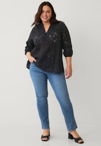 Blusa nera con bottoni, design in pizzo floreale, maniche a tre quarti e orlo leggermente curvo, abbinata a jeans dritti blu chiaro e tacchi neri.