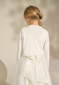 Gilet en tricot blanc avec un col en dentelle, associé à une jupe en tulle crème ornée d'un grand nœud à l'arrière.