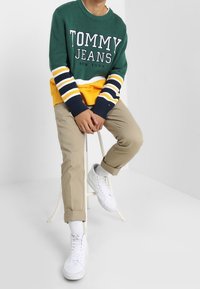 Pull en maille vert avec rayures blanches et jaunes, logo "TOMMY JEANS", pantalon kaki, baskets blanches, assis sur un tabouret devant un fond uni.